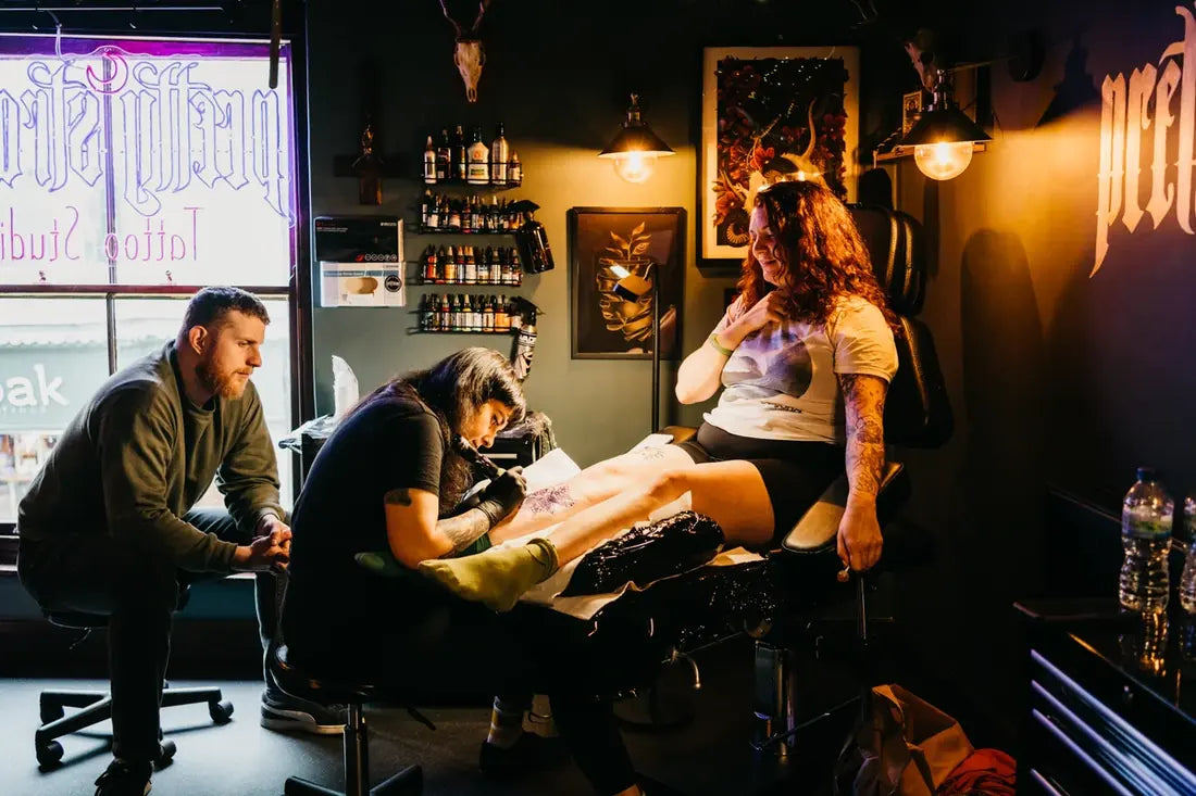 Inside Pretty Strange Tattoo Studio - an artist tattooing a client beneath the studio's window sign.
